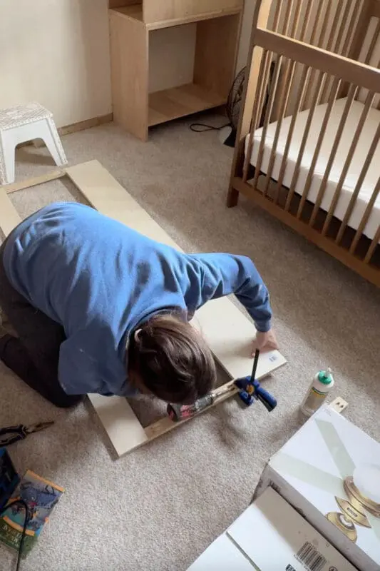 A woman assembling a wooden frame for a headboard with storage, using pocket hole screws. She is securing two long boards together with clamps and a drill while kneeling on a carpeted floor. A bottle of wood glue and other construction materials are nearby.